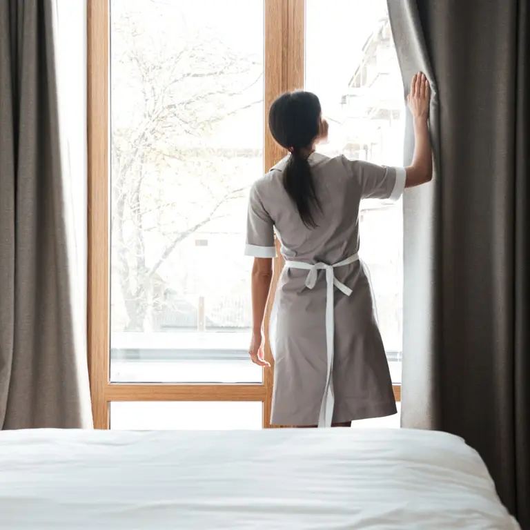 Housekeeping-Mitarbeiterin öffnet in einem hellen Hotelzimmer die Vorhänge, im Vordergrund das gemachte Bett